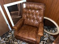 Brown leather recliner chair with tufted backrest, large rectangular white framed mirror and oval cream framed mirror positioned on patterned carpet in front of wood panel wall.