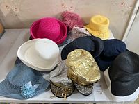 Photo showing 10 assorted summer hats including bright pink wide brim, white heart-shaped woven hat, yellow bucket hat, navy blue and black hats, sky blue hat with flower, pink knit hat, two metallic gold hats, and a scarf accessory.