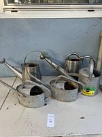 Photo showing five metal watering cans placed on a table, two small rusted galvanized cans with cut openings, and three larger metal cans with different handle and spout styles.