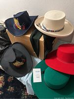 Five women's hats arranged on and around a cardboard box: two navy blue cowboy hats with dragon embroidery, one black cowboy hat, one red felt hat, and one green felt hat.
