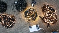 Ten coils of black wired string LED lights, several coils lit showing warm white LED bulbs, connected to power strip.