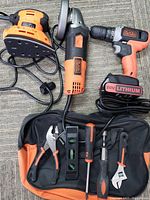 Photo of 3 Black & Decker power tools: cordless drill with 20V lithium battery, corded angle grinder, corded sander, and a black and orange bag containing hand tools including pliers, screwdrivers, spirit level.