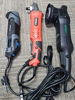 Three corded multi-tool angle grinders in different colors and brands placed side by side on carpeted floor.