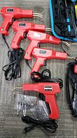 Five red plastic welding tools with power cords and assorted heads and accessories, laid out on floor.