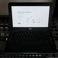 Front view of Dell Chromebook 11 3180 laptop open powered on showing welcome screen and keyboard with power supply cable visible.