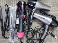 Two BaBylissPRO TT hair dryers, one black and pink round hair brush, two black and pink hair straighteners laid out showing power cords intertwined on carpet floor.