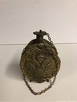 Front angle image showing the round brass flask with detailed embossed two-dragon design and attached chain to the screw cap.