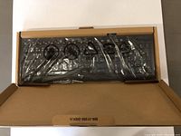 Full view of the keyboard inside an open cardboard box, covered with a clear plastic wrapping, displaying all main keys including function keys and numeric keypad.