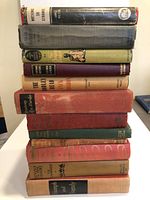 Stack of 12 hardcover historic books varying in size and color, arranged on a white table against a plain background.