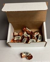 Open box containing multiple copper 90 degree elbow fittings with visible barcode labels, one fitting outside box.