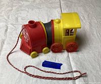 Plastic vintage train pull toy with red front, yellow cab, original red and white twisted pull string on white fabric background.