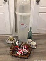 Full view of all items in lot showing basket of Christmas ribbons and decorations, tall translucent white gift wrap storage bin, green decorative bottle, serving platter, plates, and mugs on the floor.