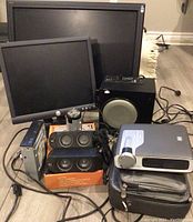 Lot of electronics showing two computer monitors, speaker system, projector, cables, and accessories.
