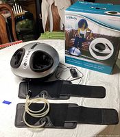 Photo of truMedic foot massager unit on table with two black fabric strap accessories and power adapter next to original box packaging showing product image and branding.