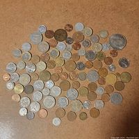 Top-down view of approximately 80 assorted world coins on a brown background, showing different sizes and colors, mostly circulated with mild to moderate wear.