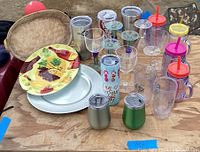 Lot of assorted used glasses, tumblers, mason jar mugs with colorful lids and straws, decorative plates with fruit motif, and metal serving platter, displayed on wood surface.
