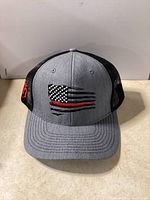 Front view of a trucker style hat with grey front, black mesh back, and embroidered American flag with red line