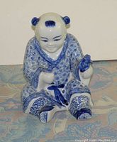 Full view of the blue and white ceramic boy figurine sitting with legs bent, detailed patterned robes visible.