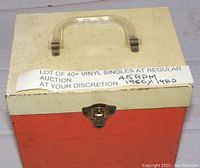Photo of a vintage red and white carrying case with a plastic handle and metal latch, labeled 'LOT OF 40+ VINYL SINGLES 45 RPM 1960/1980' on taped paper attached around the case.
