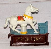 Front view of the vintage mechanical trick pony bank showing the white pony with red and gold accents on a brown base labeled 'TRICK PONY'.