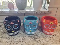 Three ceramic planters shaped and painted as Day of the Dead sugar skulls, showing black, blue, and orange/red skull planters with flowers around the eyes and mouth teeth pattern, placed on a countertop beneath a window.
