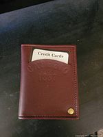 Photo of the front of a brown leather bi-fold wallet with embossed 'Capezio 1887' and a brass rivet at the bottom corner on a black background.