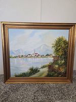 Framed oil painting showing a peaceful European Bavarian village scene with lake, village houses, distant mountains, and trees along the shore.
