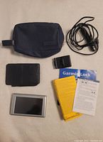Photo showing Garmin GPS device, black power cable, mounting bracket, carrying pouch, and printed manuals including Garmin Lock card and user guides.