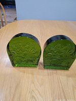 Two solid translucent green glass bookends with embossed floral or vine patterns positioned on a wooden table.