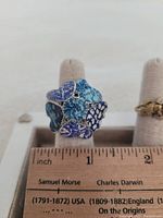 Photo of a large blue flower-shaped costume jewelry ring on a beige ring holder. The ring rests next to a ruler showing its approximate size of about 1.5 inches diameter.