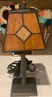Full view of a small stained glass table lamp showing its orange geometric shade and gray metal base with cord coiled at its back.