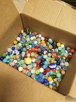 Photo of a cardboard box filled with a large quantity of vintage glass marbles of assorted colors and patterns.