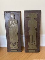 Front view of two wooden plaques with mounted brass figurative figures in medieval style.