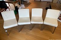 Four white vinyl upholstered dining chairs with metal legs arranged in a row on a wooden floor indoors.