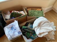 Three cardboard boxes filled with assorted folded tablecloths, napkins, and placemats with different colors and patterns; a white crocheted or lace piece of fabric partially unfolded on the floor next to the boxes
