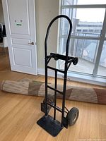Front side view of the black metal convertible hand truck dolly with a folded base plate and handle upright, showing two pneumatic tires and a curved handle.