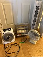 Full frontal view of the four items: tower fan, desk fan, metal heater, and box fan heater on wooden floor against white doors.