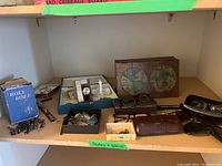 Overview of lot contents on wooden shelf: blue Holy Bible, black bead rosary, two boxes of pins and cufflinks, eyeglass frames, wooden decorative box at back, green jewelry box