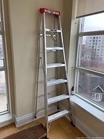 Pro Lite 7-step aluminum ladder standing indoors near windows with wooden flooring visible.