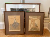 Three framed lithograph prints leaning against a wall. One large black and white lithograph, two smaller colored lithographs.