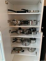 Set of pots and pans stored on five shelves in a white cabinet including frying pans, saucepans, strainer, and steamer insert.