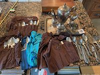 Set of silver plate cutlery rolled in cloth wraps beside silver tray and tea set on countertop.