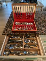 Open wooden case with red lining showing silver plated cutlery including knives, forks, spoons, and serving utensils arranged neatly.