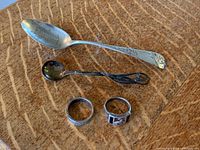 Photo showing the two silver rings and two silver spoons laid flat, highlighting the engraving on the larger spoon and the design on the smaller spoon and rings.