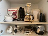 Shelved collection showing multiple small kitchen appliances including coffee maker, kettle, blender, and hand mixers.