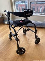 Full side view of black Nexus rollator mobility walker showing cushioned seat and backrest strap in a room with wooden floor and windows in background.