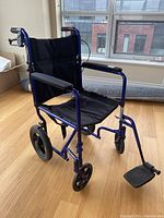 Front-right angled view of the Invacare manual wheelchair showing blue metal frame, black fabric seat and back, front caster wheels, and left footrest. Missing right footrest visible.