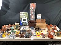 Overview of table with assorted firearm accessories including holsters, grips, scope mounts, bipod, slings, shooting bags, cleaning supplies