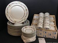 Stack of twelve 9″ dessert plates with floral and gold trim pattern alongside wrapped stacks of saucers and carton of demitasse cups