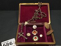 Overview of the collection inside the open men's jewelry box, showing rosary, pins, and flat pennies on burgundy fabric lining.